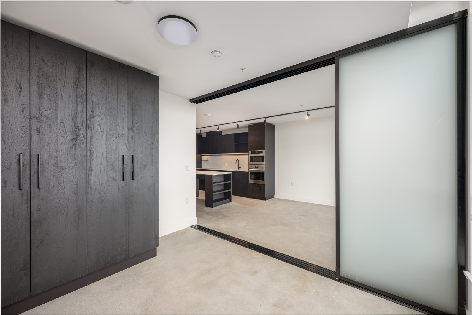 A modern apartment with dark wood cabinets, a sliding frosted glass partition, concrete floors, and an open kitchen with built-in appliances.