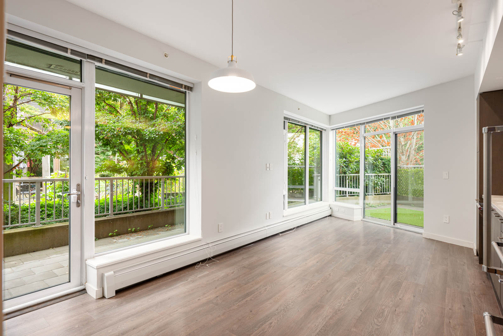 Empty, unfurnished room with large floor-to-ceiling windows and glass doors opening to a patio, featuring wood flooring and a white pendant ceiling light.