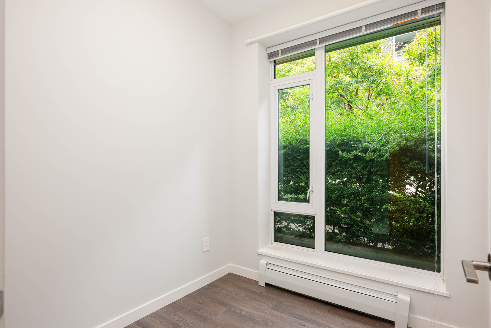 Small empty room with white walls, large window, brown hardwood floor, and view of green trees outside.