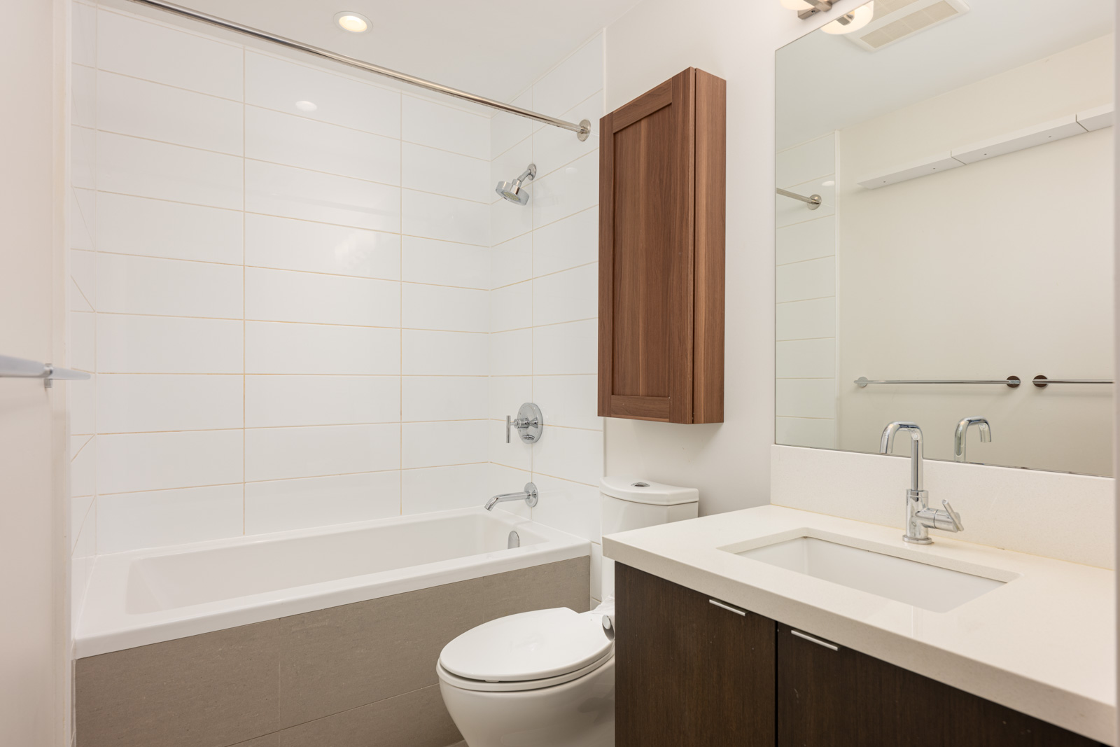 Modern bathroom with white tile bathtub-shower combo, wall-mounted wooden cabinet, toilet, and white countertop sink with chrome fixtures under a large mirror.