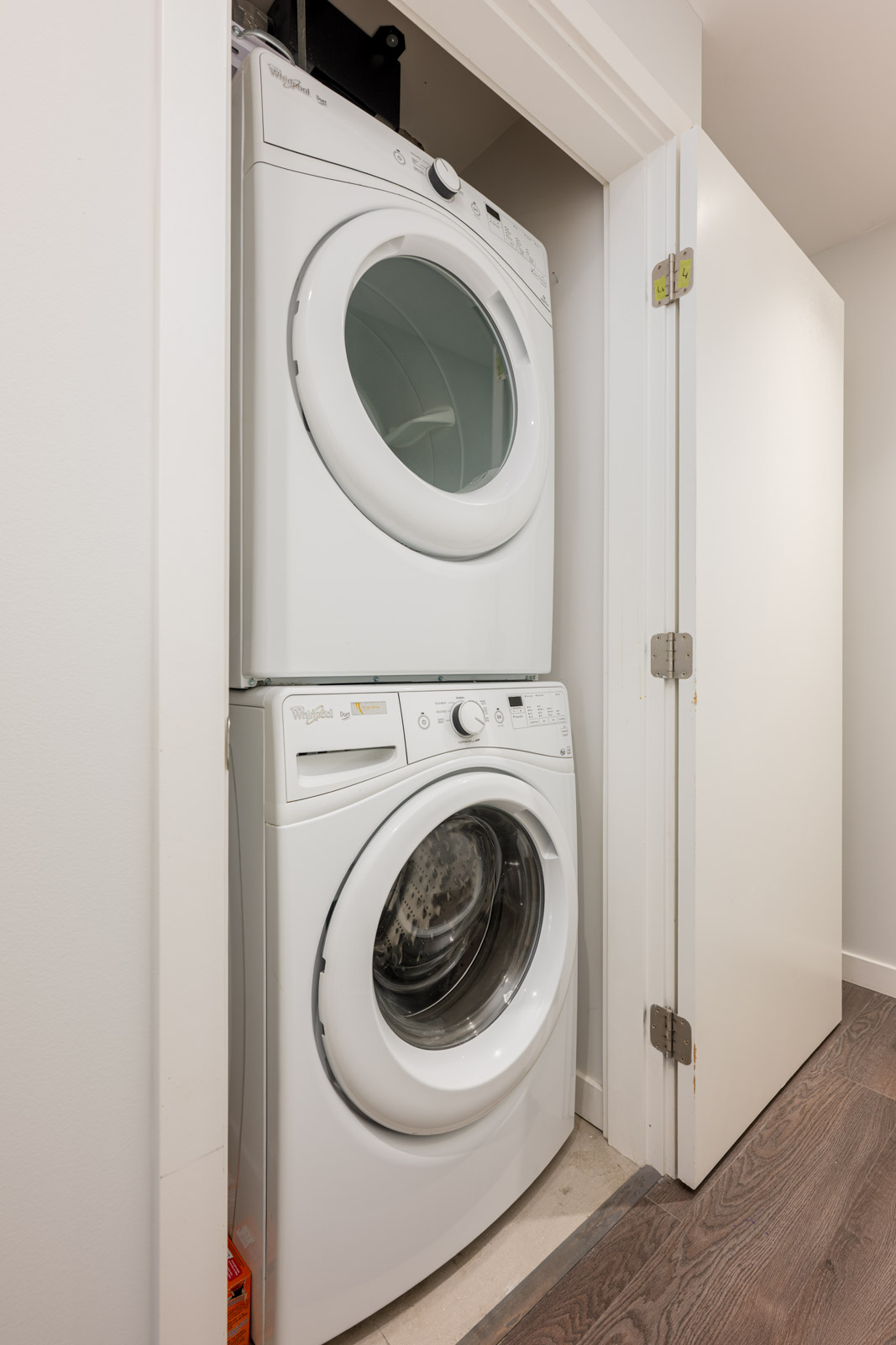 Stacked front-loading washer and dryer units in a small laundry closet with a white door open, showing the machines against a white wall and wood flooring.