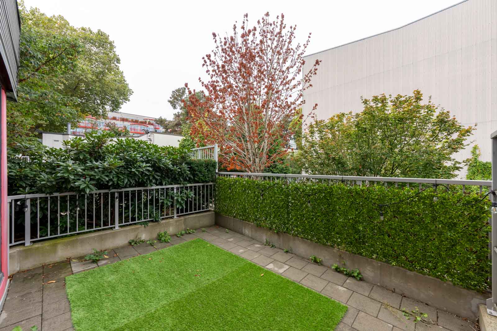Small outdoor patio with artificial grass, surrounded by metal railings and green hedges, adjacent to trees and buildings in the background.
