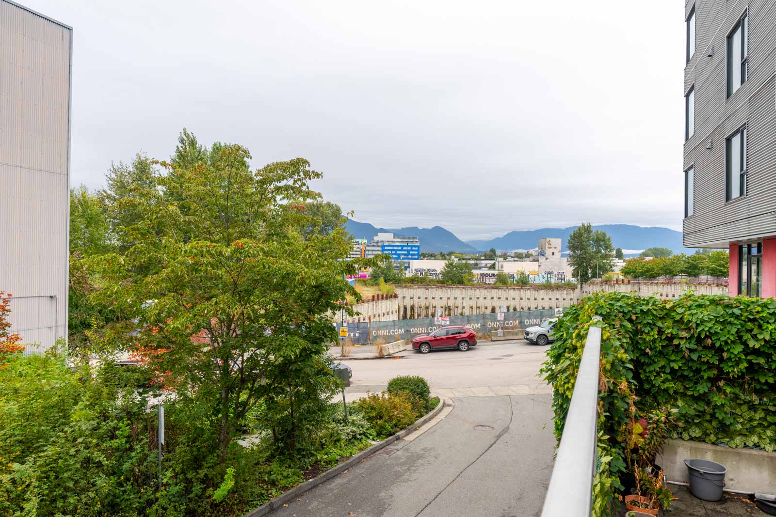 A construction site with wooden pilings is visible behind trees and parked cars; buildings and mountains are in the background under an overcast sky.