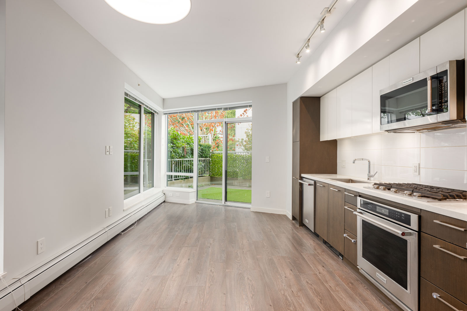 Modern kitchen with stainless steel appliances and dark cabinets, adjacent to an empty living area with large windows and glass doors opening to a patio with greenery outside.
