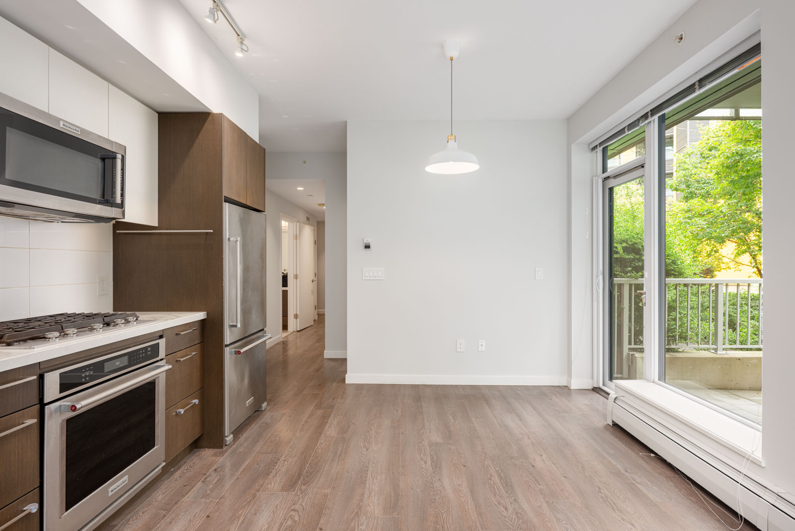 Modern kitchen with stainless steel appliances, wood cabinets, and large window leading to a patio; features light wood flooring and white walls.