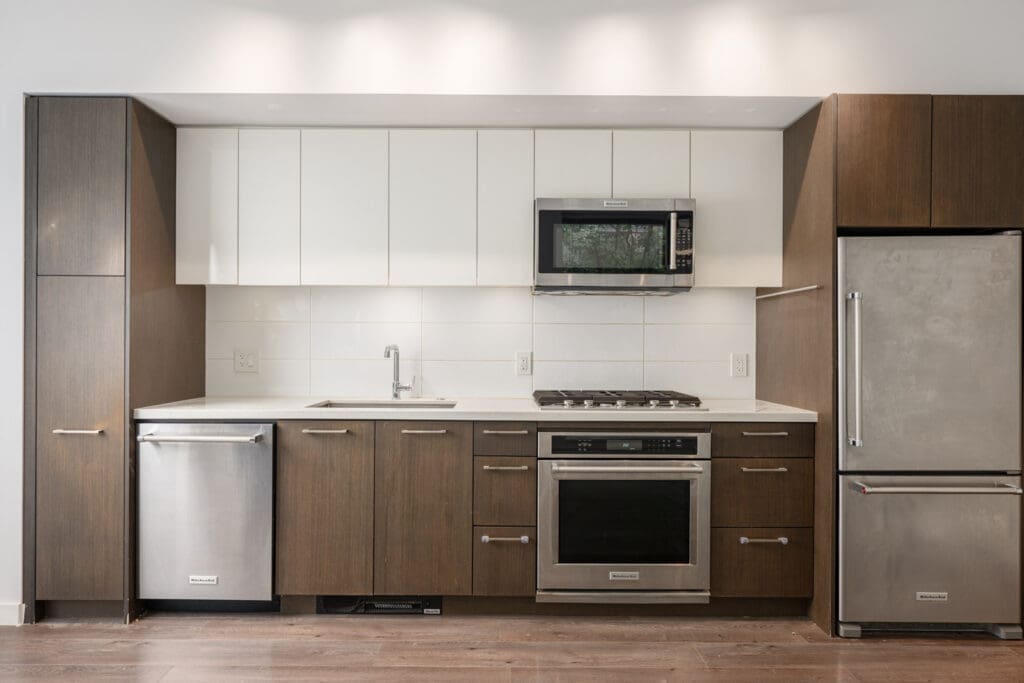 Modern kitchen with brown cabinets, white upper cabinets, stainless steel appliances, and a white tile backsplash. Includes a sink, dishwasher, oven, microwave, stovetop, and refrigerator.