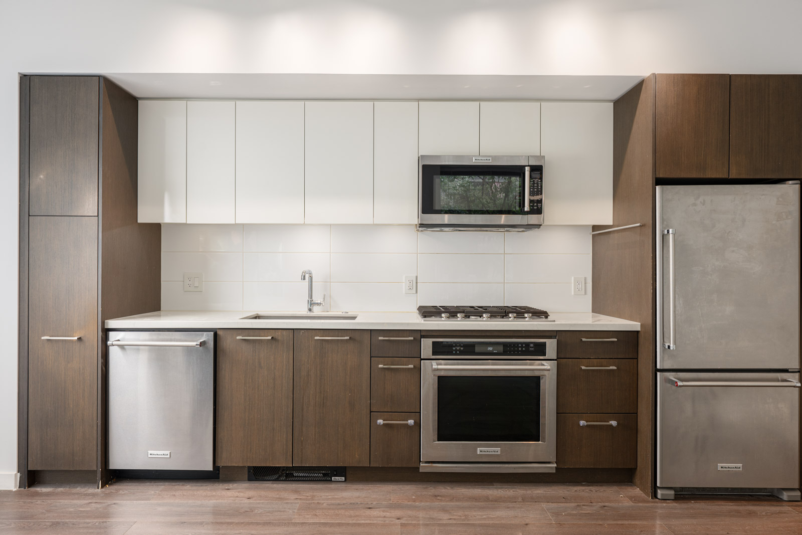 Modern kitchen with brown cabinets, white upper cabinets, stainless steel appliances, and a white tile backsplash. Includes a sink, dishwasher, oven, microwave, stovetop, and refrigerator.