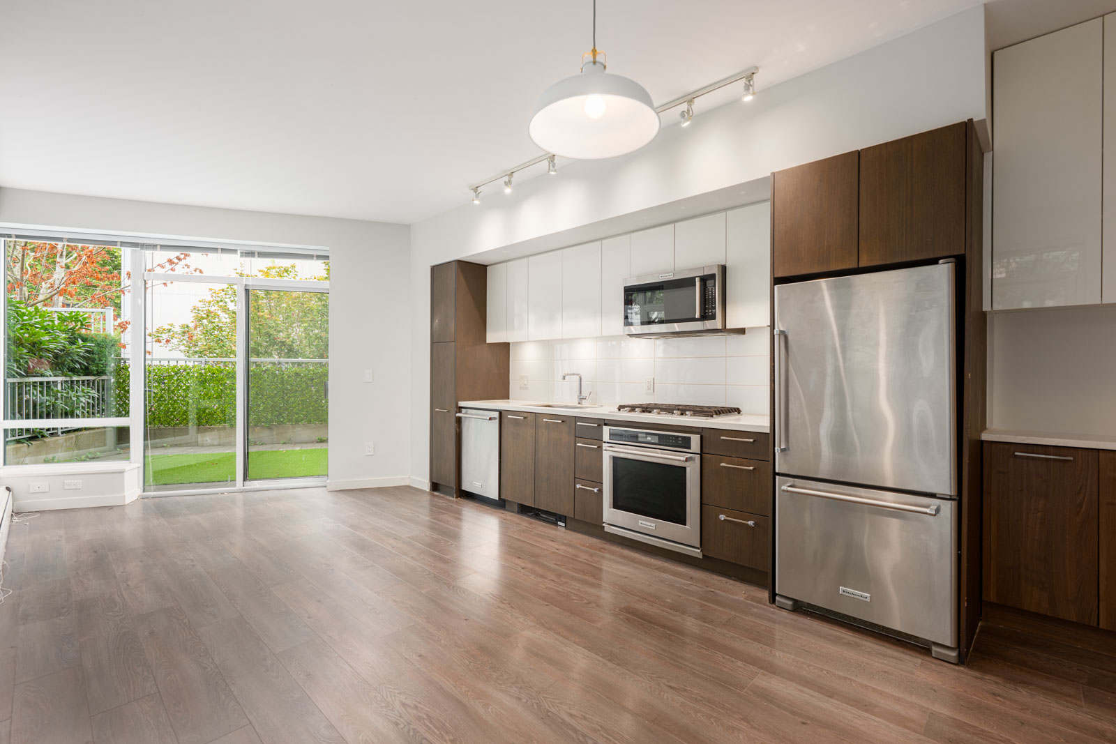 Modern kitchen with stainless steel appliances, dark wood cabinets, white backsplash, and light wood flooring, adjacent to large windows overlooking a green outdoor area.