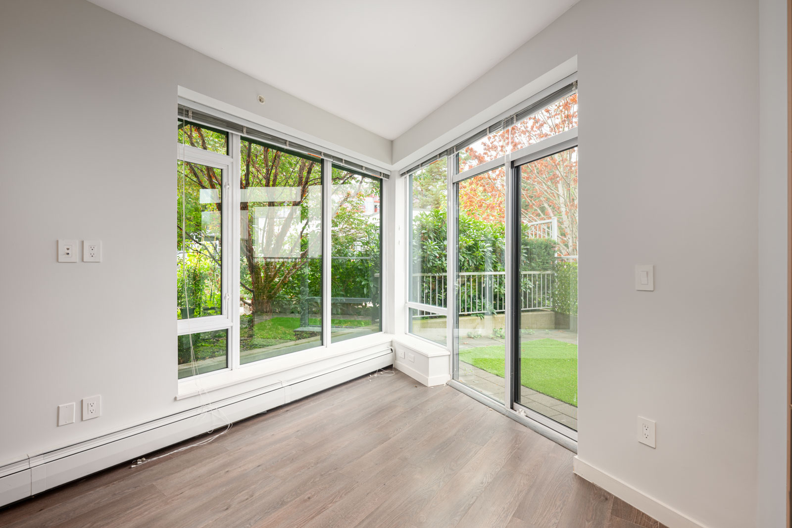 Empty room with light gray walls, large windows, and sliding glass doors opening to a small patio with green grass and trees outside.
