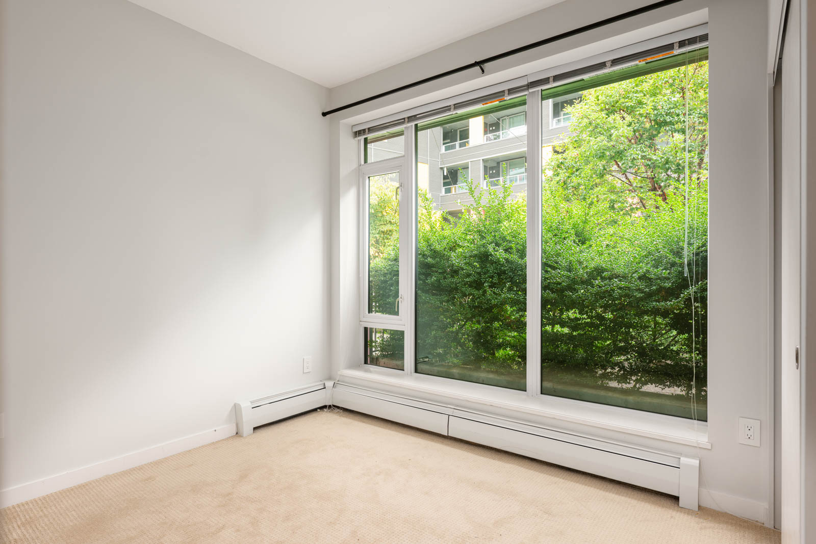 Empty room with beige carpet, white walls, and a large window overlooking green bushes and an apartment building outside.
