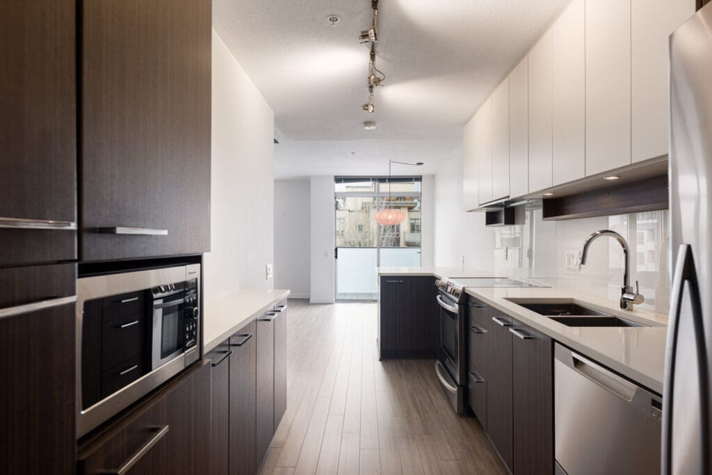 Modern galley kitchen with dark and white cabinets, stainless steel appliances, quartz countertops, and wood flooring, leading to a balcony with glass doors.
