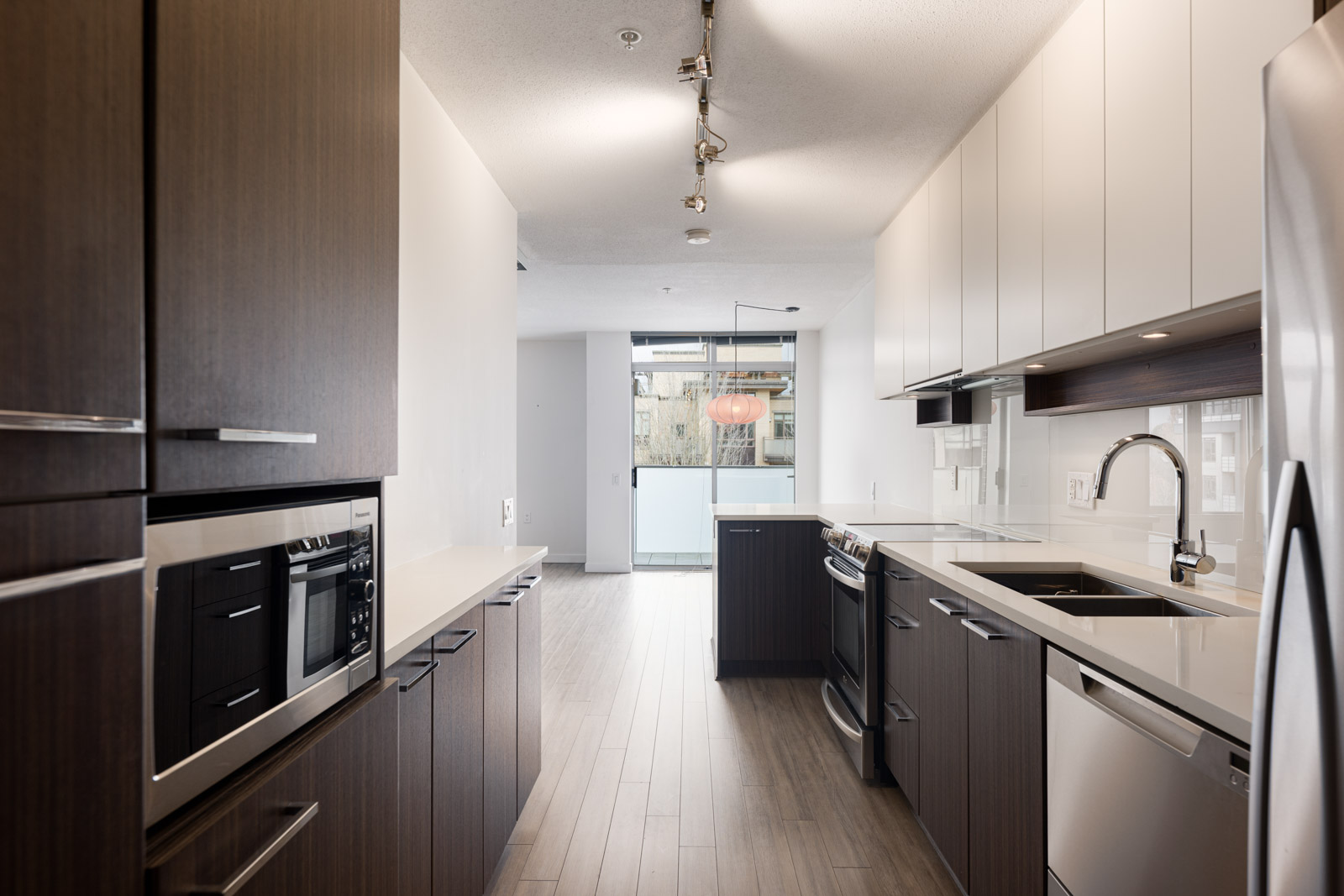Modern galley kitchen with dark and white cabinets, stainless steel appliances, quartz countertops, and wood flooring, leading to a balcony with glass doors.