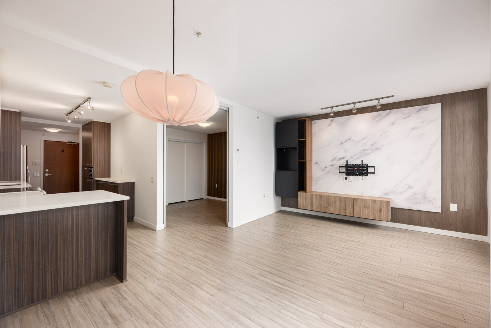 Modern unfurnished living room with wood flooring, a marble accent wall with mounted TV bracket, built-in cabinets, and an open kitchen area with dark wood cabinetry.