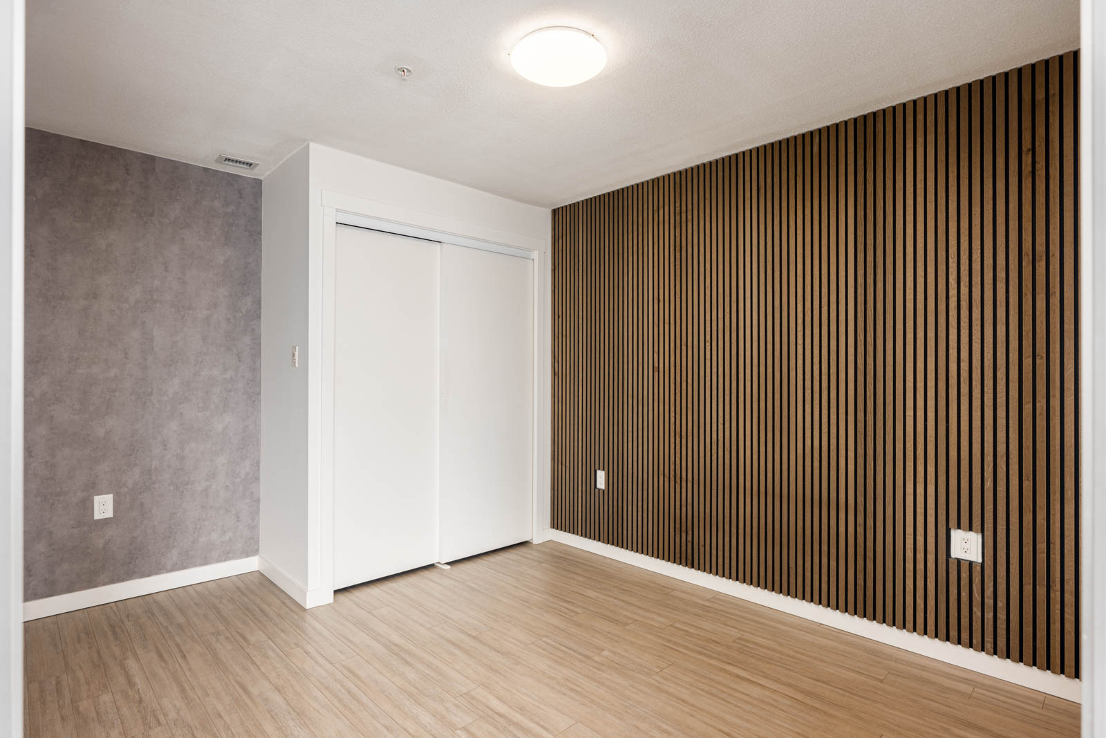 Empty room with wood-patterned floor, a slatted wood accent wall on the right, gray wall on the left, and double white sliding closet doors. Ceiling has a round light fixture.