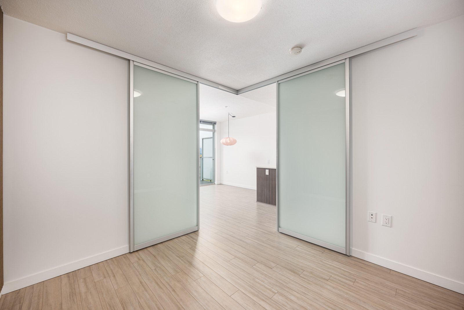 A room with light wood flooring and frosted glass sliding doors opens to a modern, bright living area with a hanging light fixture.
