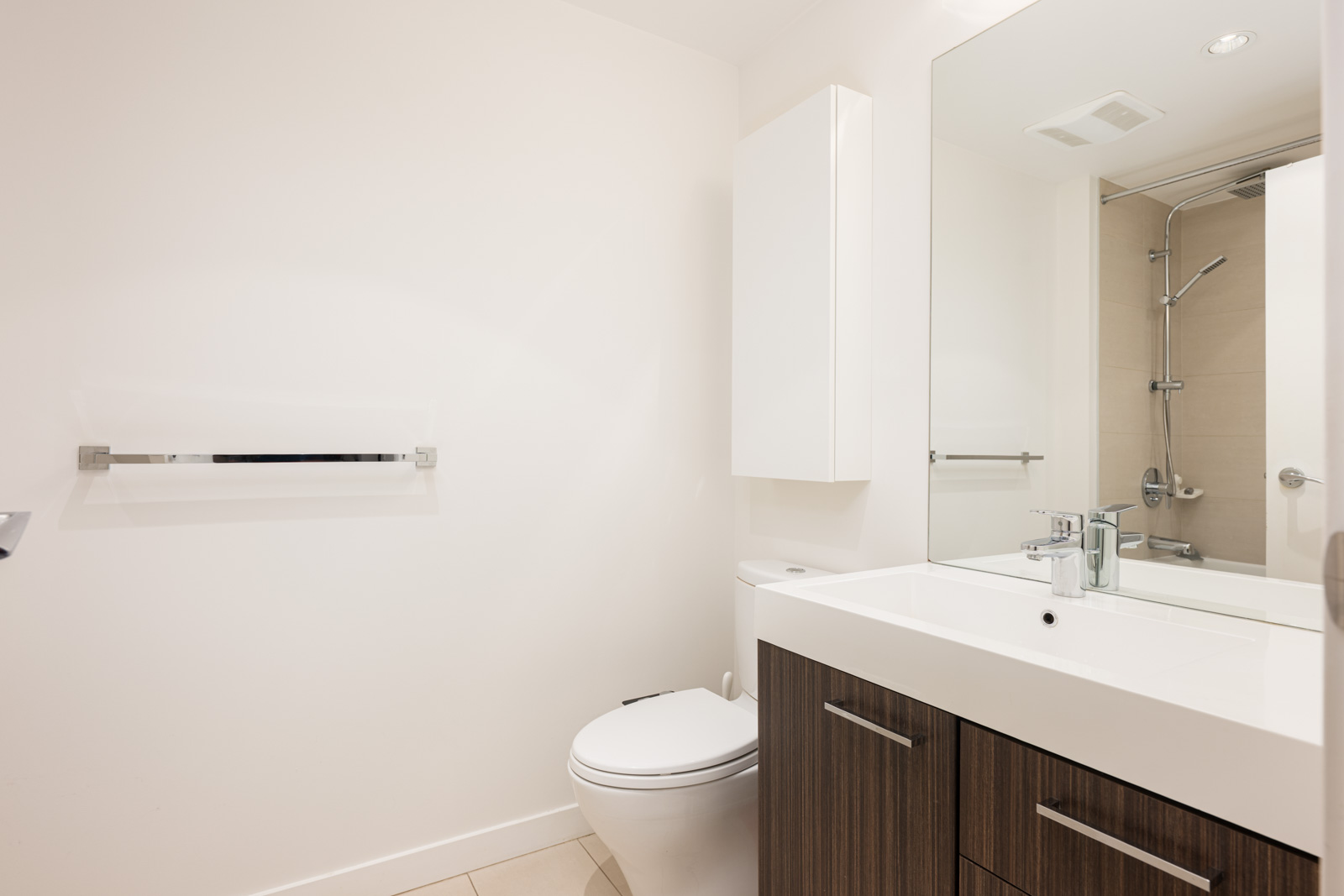 Modern bathroom with white walls, a toilet, a wall-mounted towel rack, a large mirror, a sink with dark wood cabinetry, and a glass-enclosed shower.