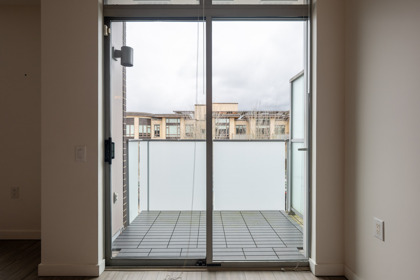 A glass sliding door opens to a small balcony with frosted glass panels, overlooking neighboring buildings under a cloudy sky.