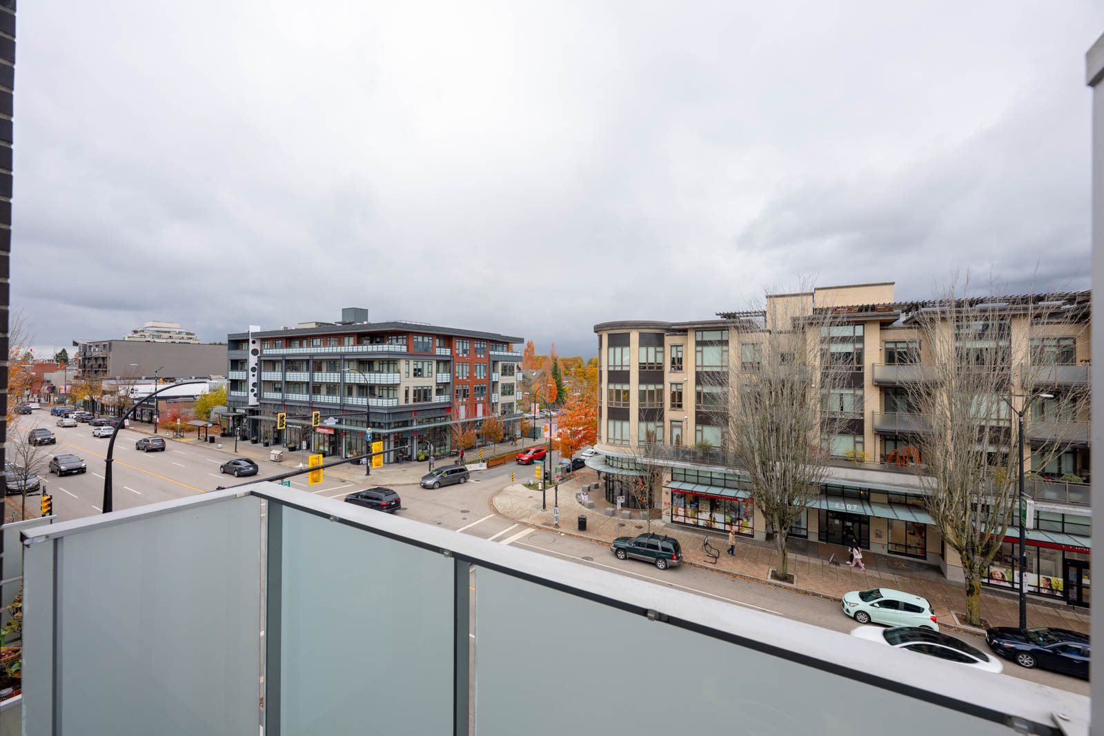 View from a balcony overlooking a busy urban intersection with modern apartment buildings, stores, cars, and overcast sky.