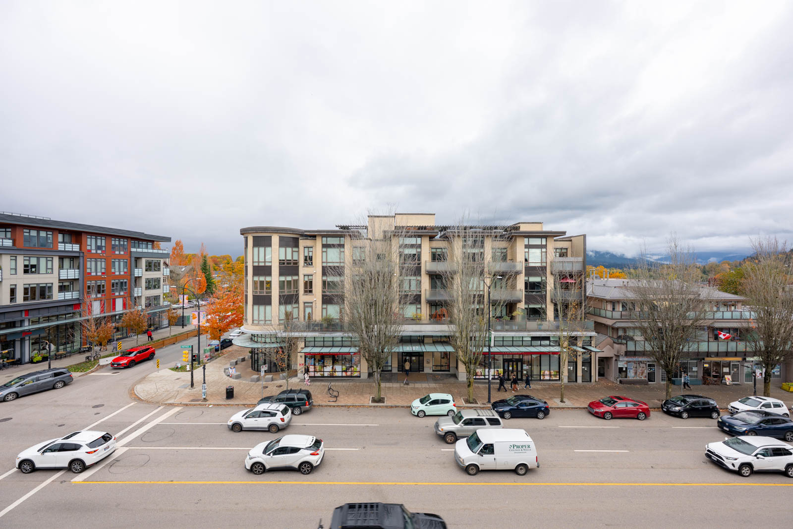 A four-story mixed-use building with shops at street level, parked cars on the curb, and moving traffic on a cloudy day. Trees in front have lost most of their leaves.