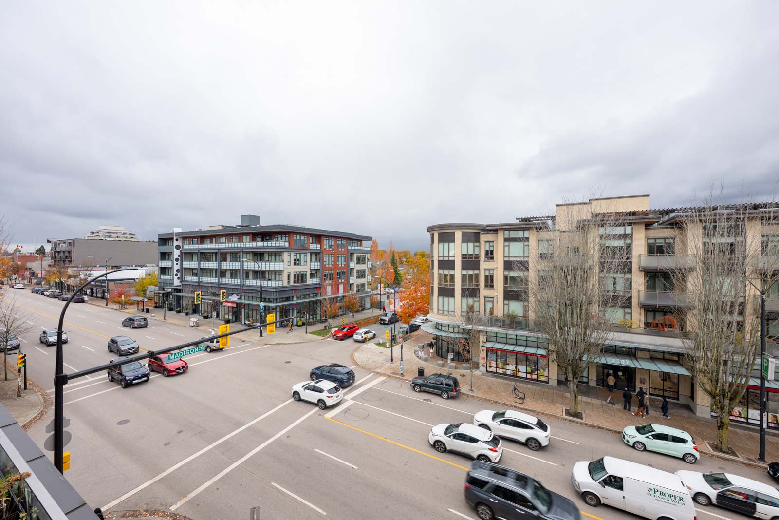 A wide intersection in a city with cars driving and parked, pedestrians on sidewalks, and modern multi-story buildings under a cloudy sky.