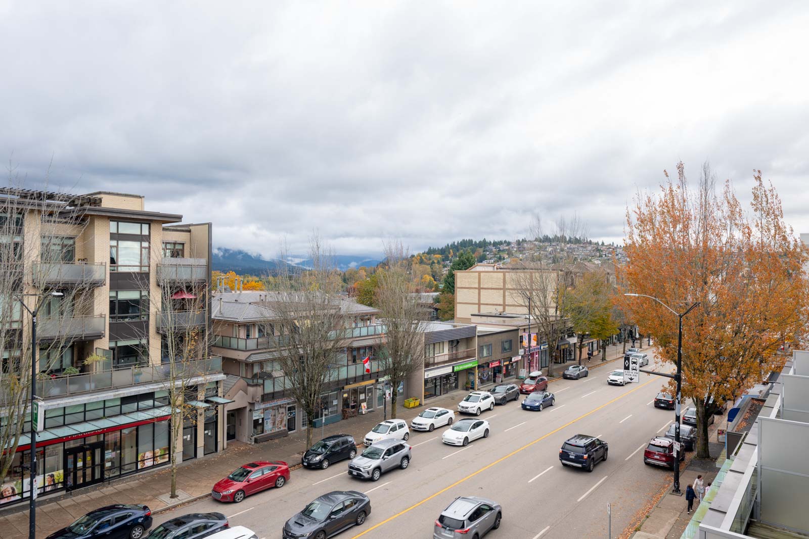 A city street with cars driving and parked along both sides, lined with low-rise buildings and trees, under a cloudy sky. Hills are visible in the background.