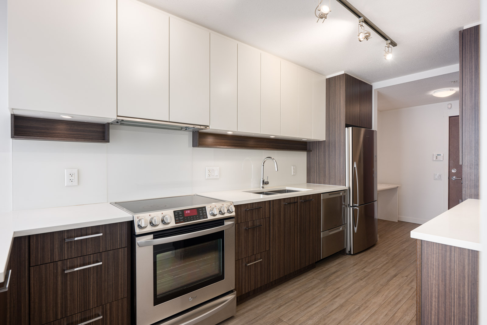 Modern kitchen with stainless steel appliances, light and dark wood cabinets, electric stove, sink, refrigerator, and overhead track lighting on wood-patterned flooring.