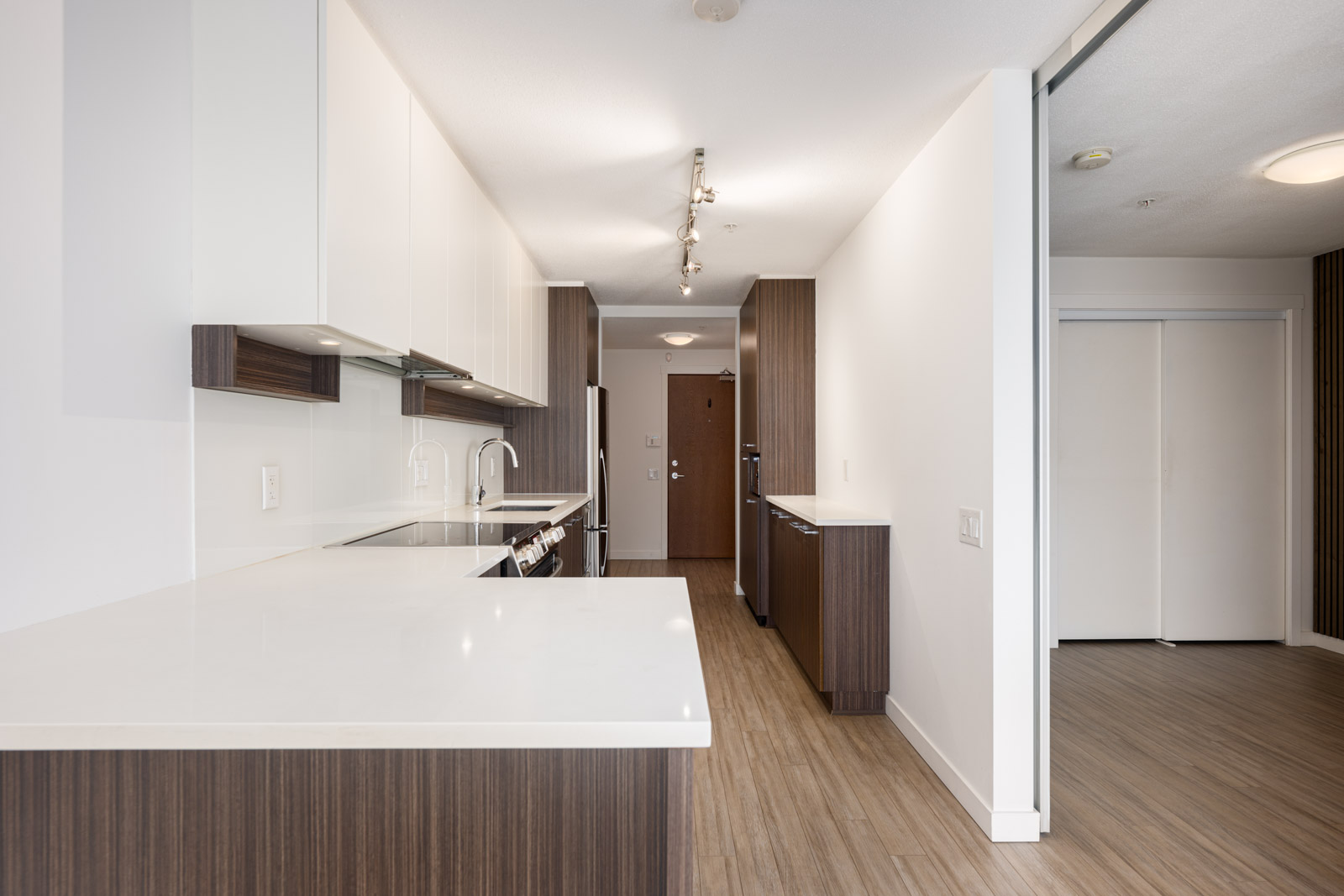Modern kitchen with white countertops, wood cabinetry, stainless steel appliances, and track lighting, leading to a hallway with wooden floors and a closed door at the end.