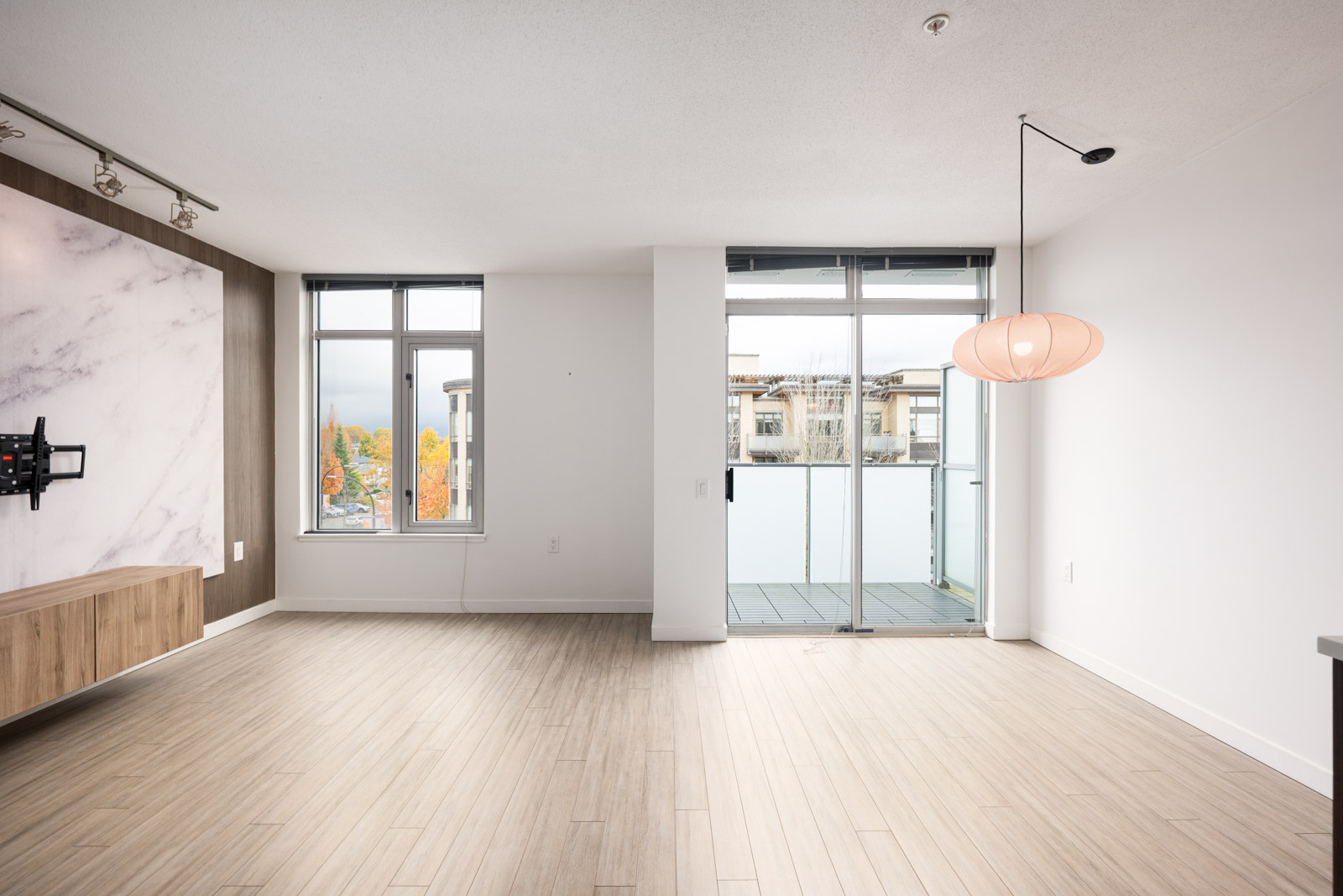 Unfurnished modern living room with light wood floors, a mounted TV bracket, large windows, balcony access, and a hanging light fixture.