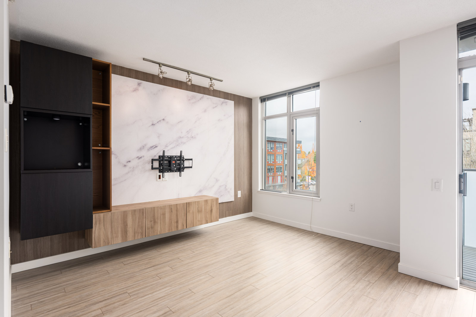 Modern, unfurnished living room with a wall-mounted TV bracket, wood and marble accent wall, light wood flooring, large window, and sliding glass door.