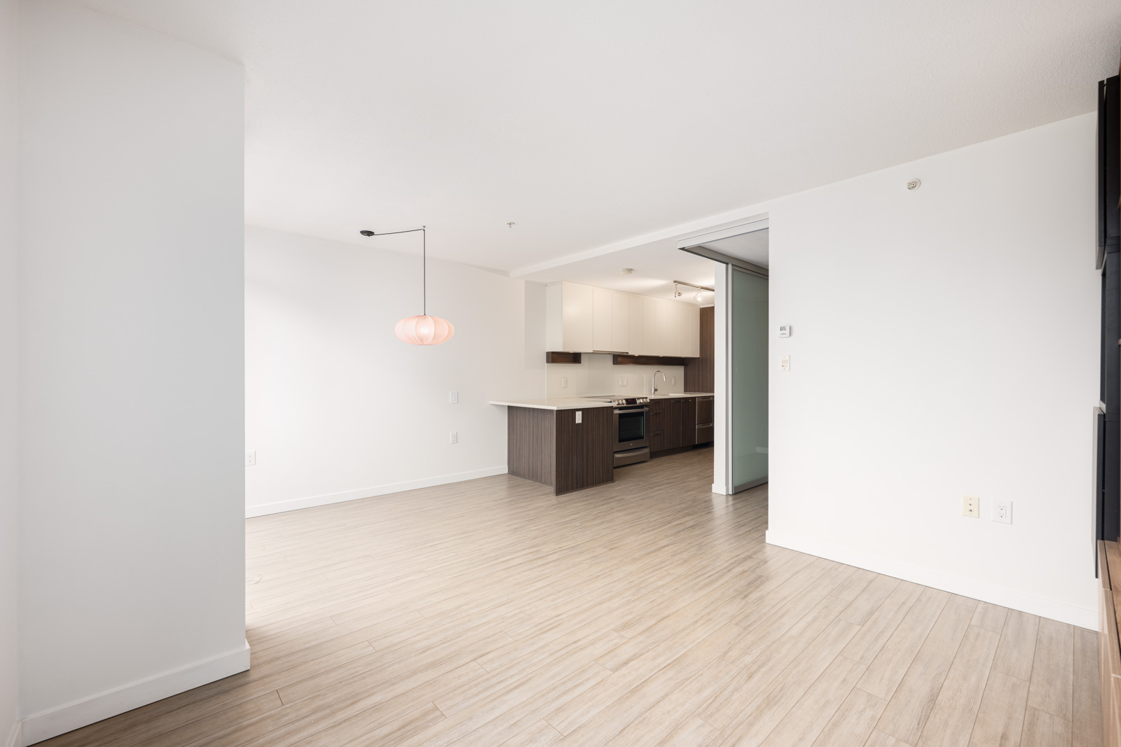 Unfurnished modern apartment interior with light wood flooring, white walls, open kitchen area, and hanging pendant light over the dining space.