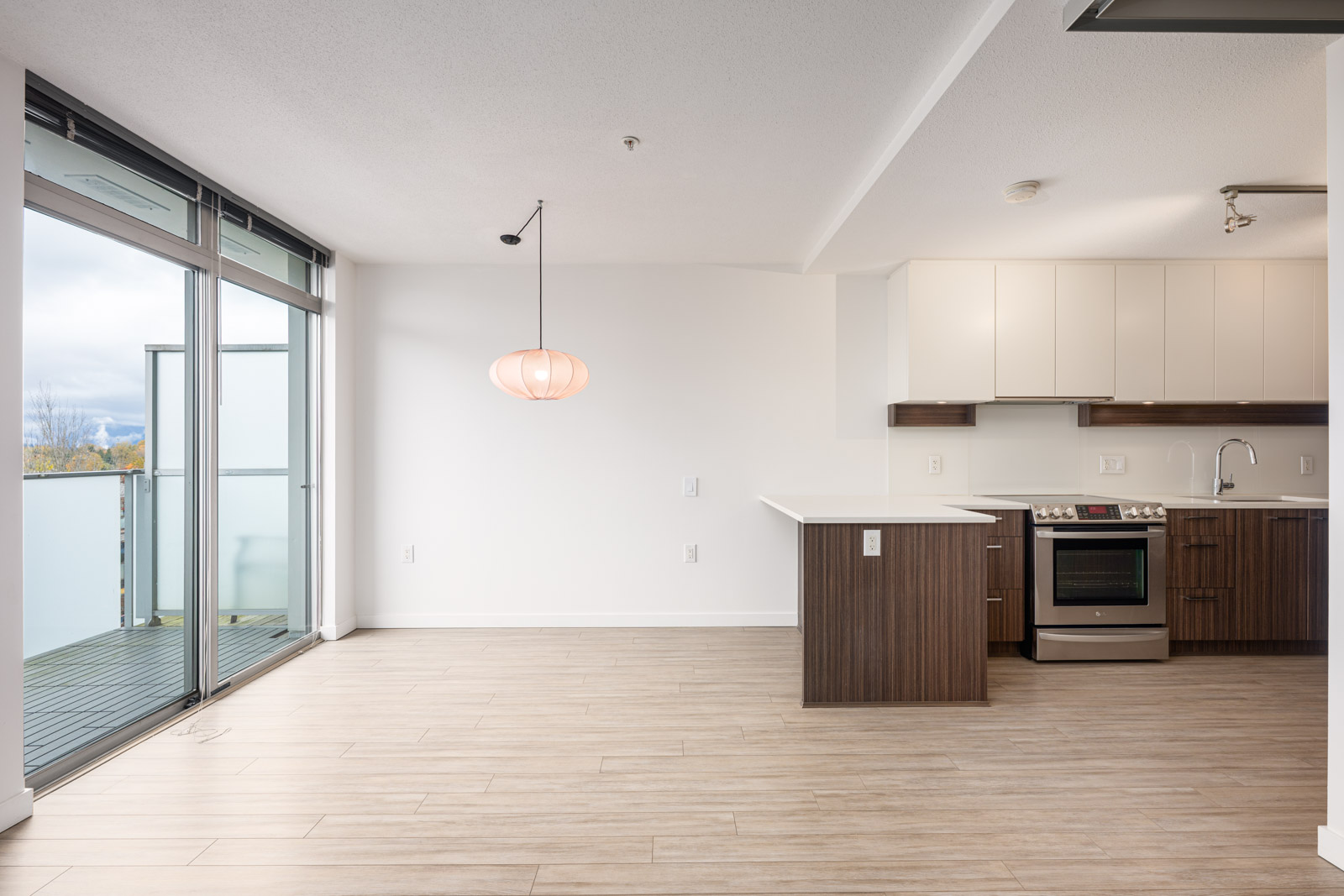 Modern, unfurnished apartment kitchen and dining area with wood floors, white walls, built-in cabinets, stainless steel appliances, and large windows leading to a balcony.
