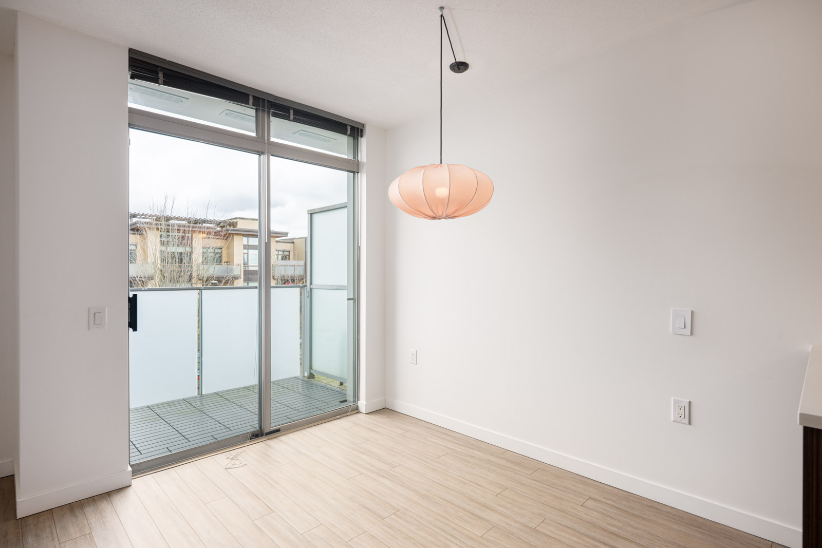 Empty room with light wood floor, white walls, a modern pendant light, and a glass door leading to a small balcony with frosted panels.