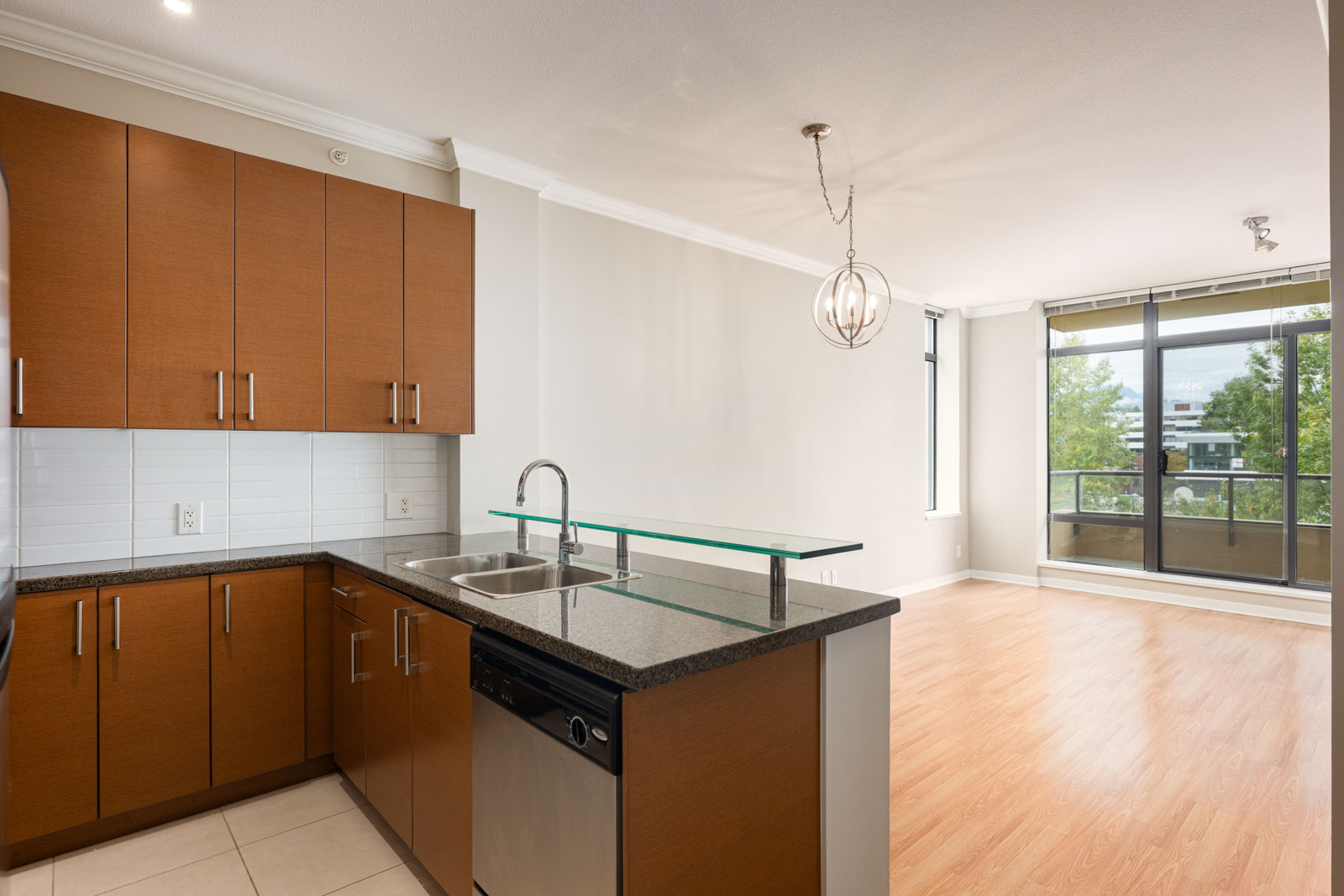 Modern apartment kitchen with brown cabinets, black countertops, and stainless steel appliances, open to a bright living area with large windows and wood flooring.