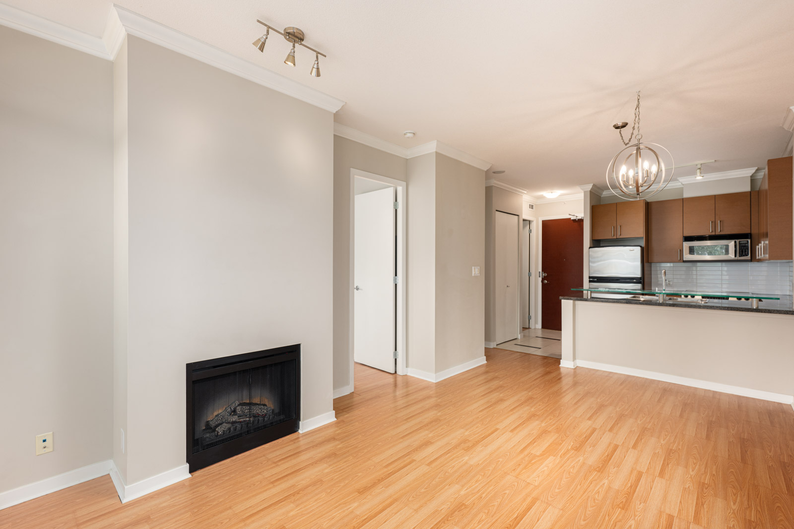 Unfurnished apartment interior with light wood flooring, a fireplace, neutral walls, and a modern kitchen featuring stainless steel appliances and pendant lighting.