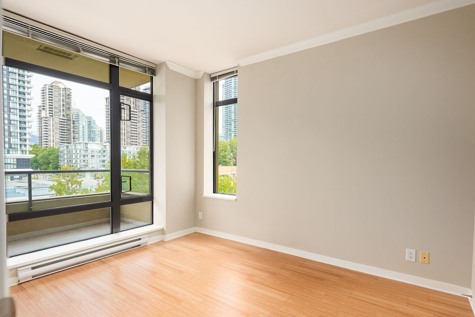 Empty room with light wood flooring, beige walls, and large windows showing views of nearby modern high-rise buildings and greenery.