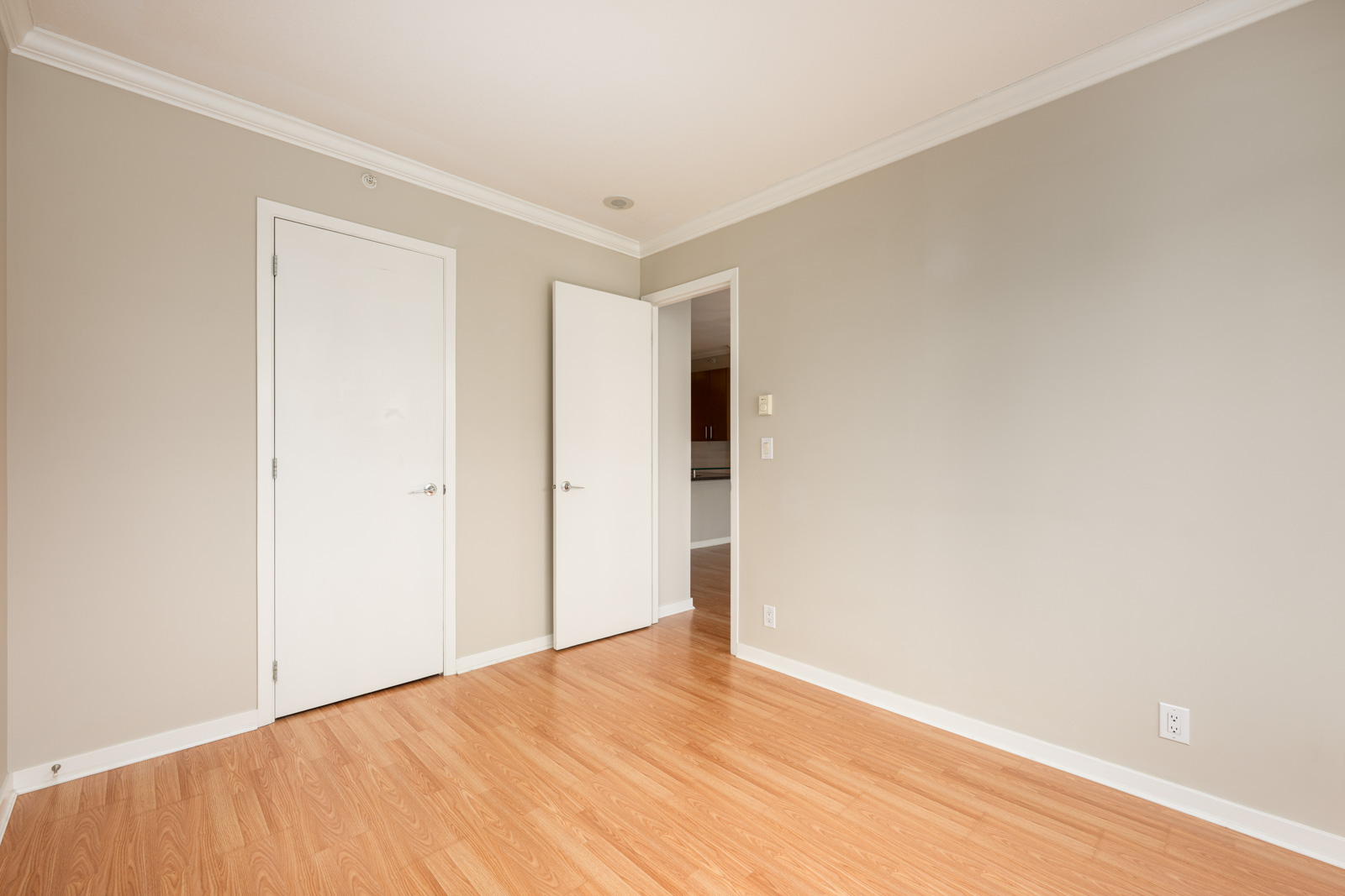 Empty room with light wood flooring, beige walls, white trim, two closed white doors, and one open door leading to another area.