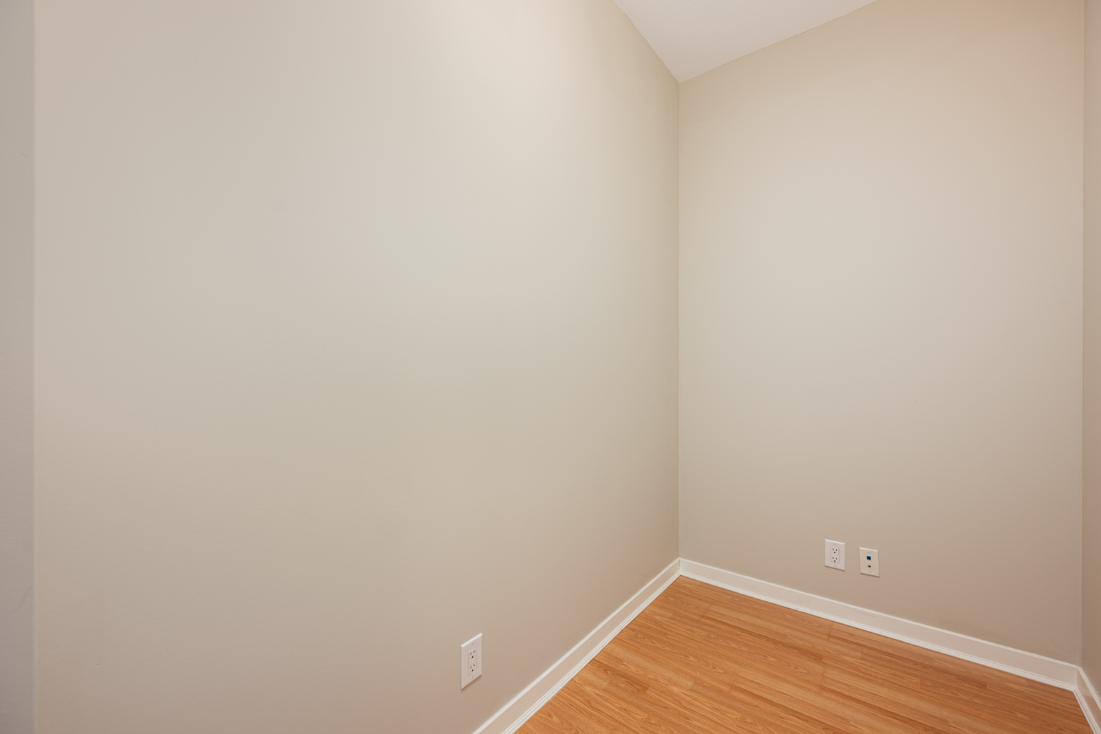 Empty corner of a small room with beige walls, white baseboard, light wood floor, and electrical outlets on the wall.