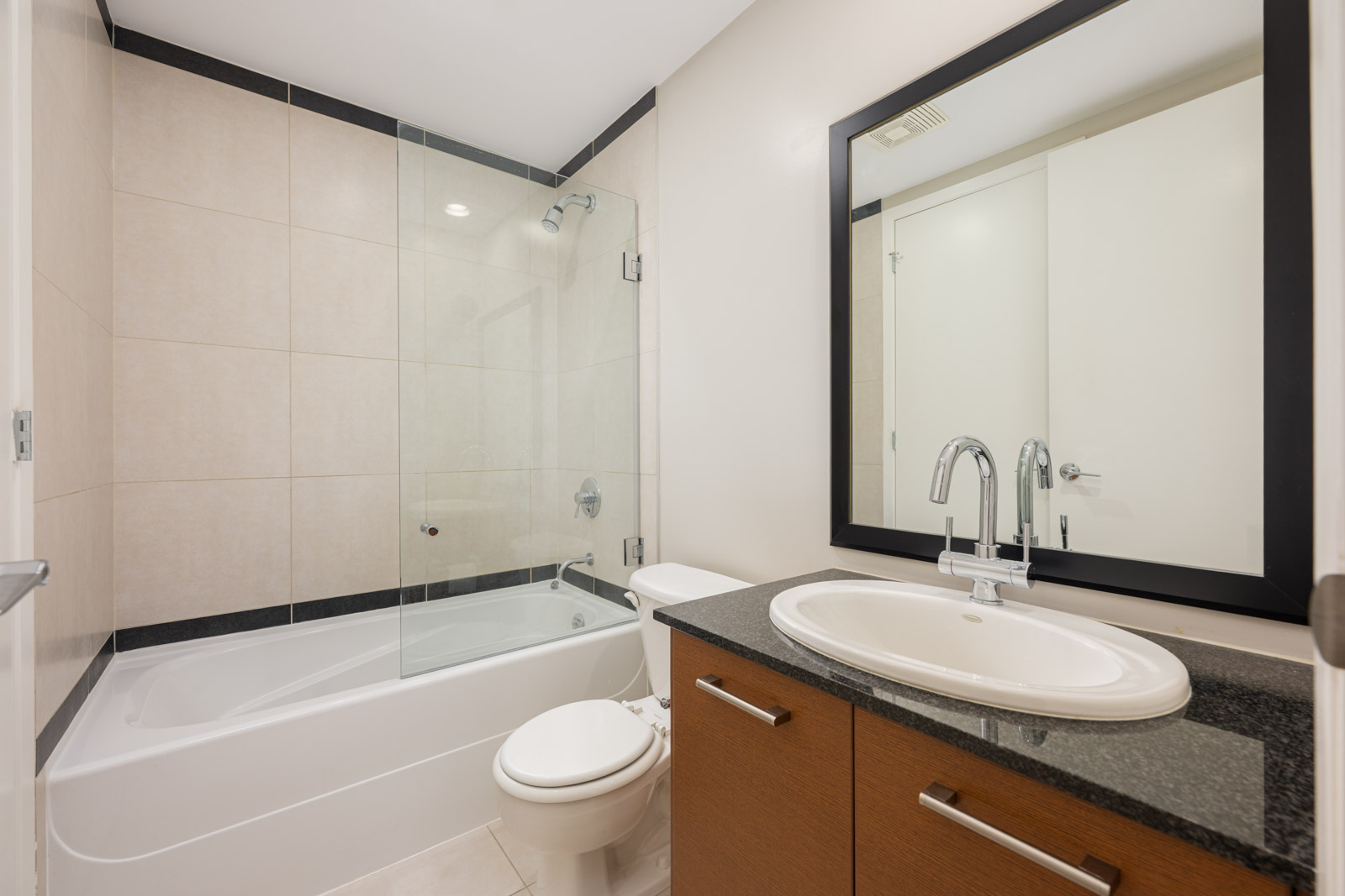Modern bathroom with bathtub-shower combo, glass partition, toilet, and sink with dark countertop, mirror, and wooden cabinet.