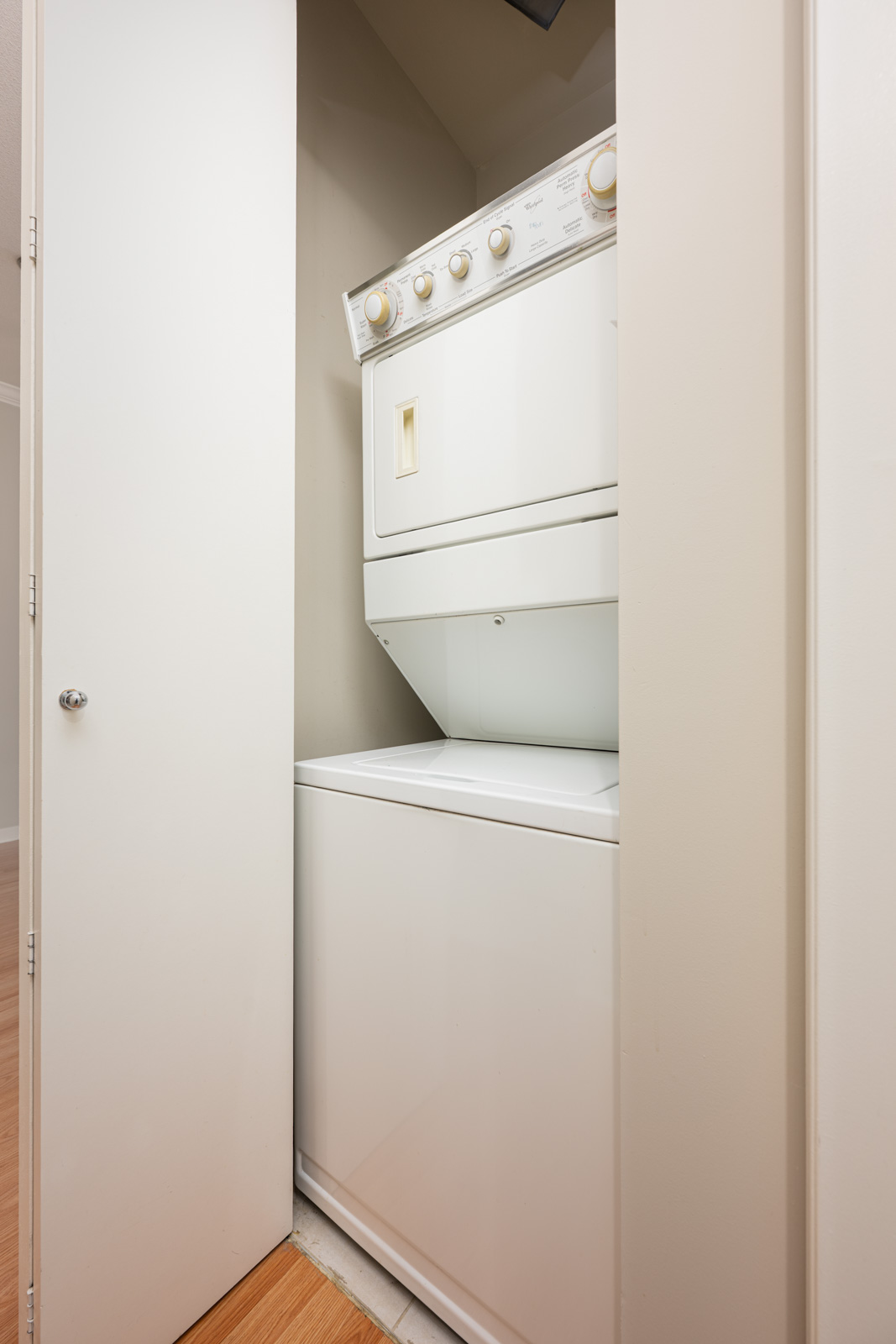A white stacked washer and dryer unit is enclosed in a small laundry closet with the door partially open.