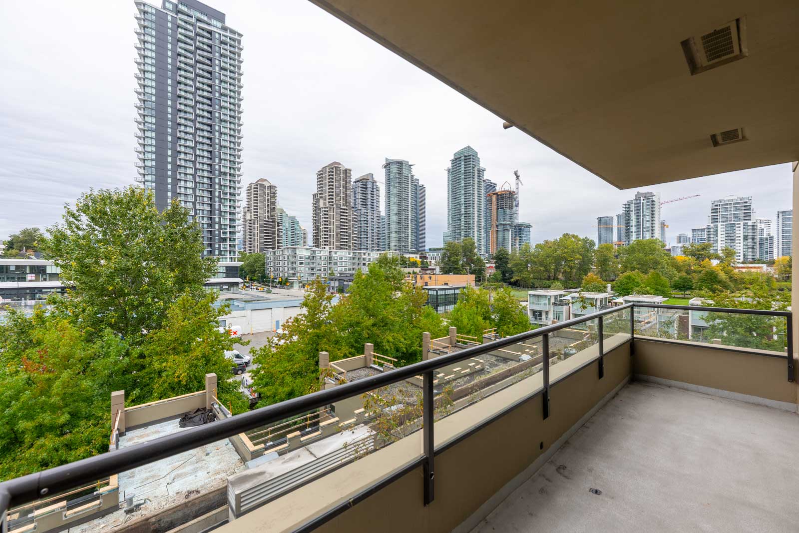 View from a balcony showing glass railing, leafy trees below, and several high-rise buildings under a cloudy sky in the background.