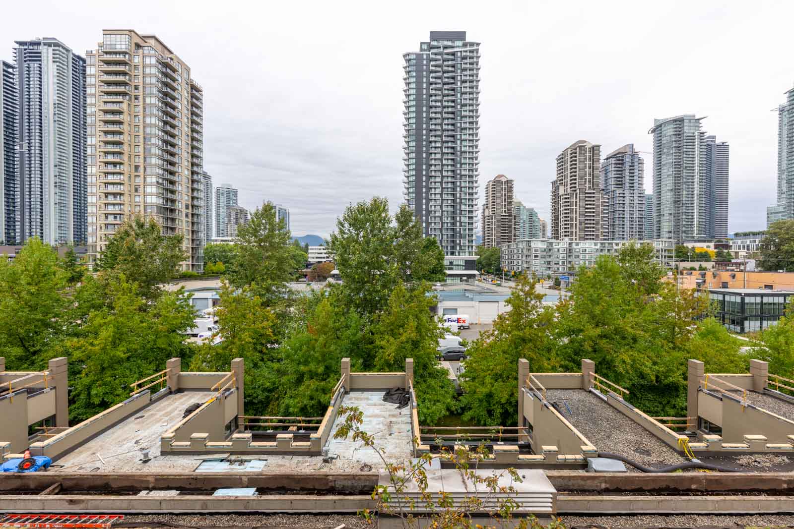 Tall modern apartment buildings and high-rises stand behind a row of green trees and rooftops on an overcast day in an urban cityscape.