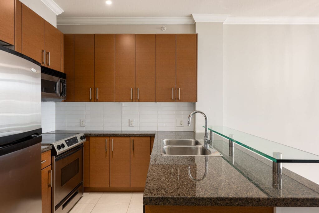 Modern kitchen with brown cabinets, stainless steel appliances, granite countertops, double sink, and a raised glass bar, with white tiled backsplash and beige walls.