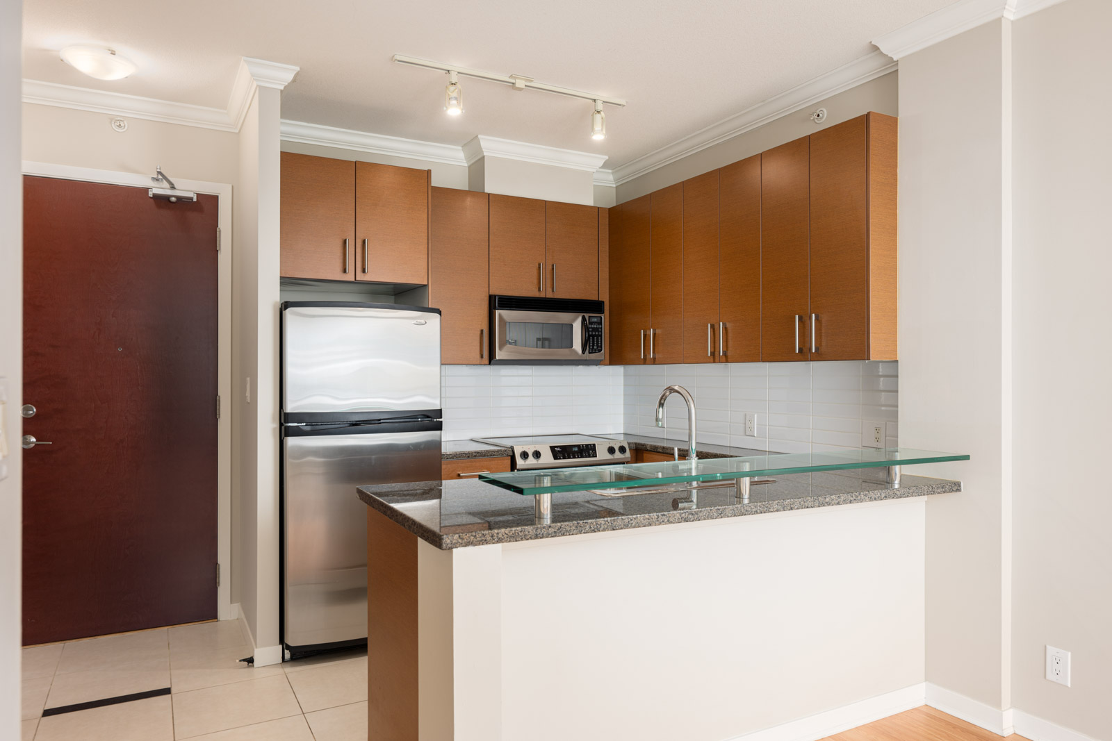 Modern kitchen with wooden cabinets, stainless steel appliances, granite countertop, and a glass panel on the island; white tile floor and backsplash.