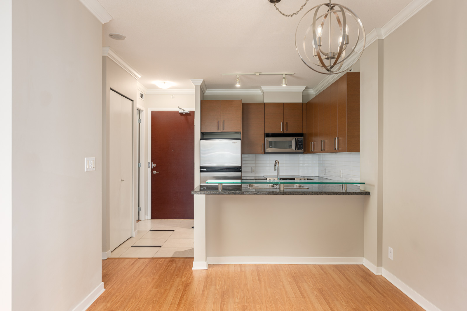 Modern kitchen with wood cabinets, stainless steel appliances, granite countertop, and a glass pendant light. Entry door and tile flooring visible in the background.