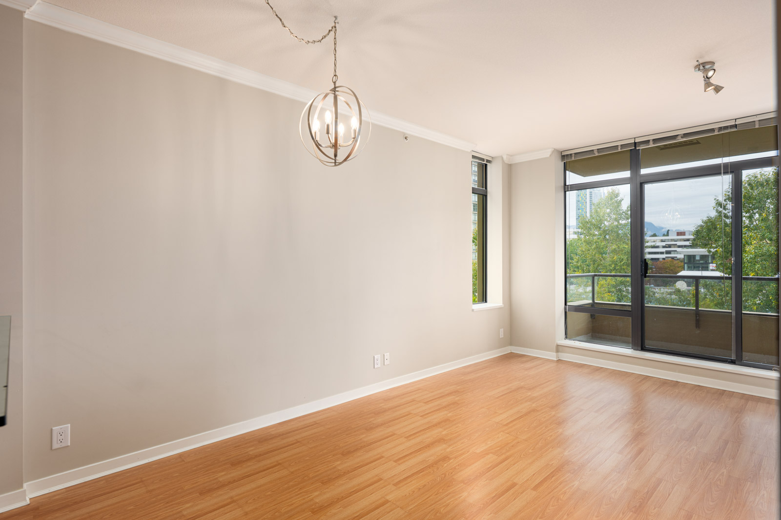 Empty room with light wood flooring, beige walls, a modern ceiling light fixture, and large windows leading to a balcony with outdoor views.