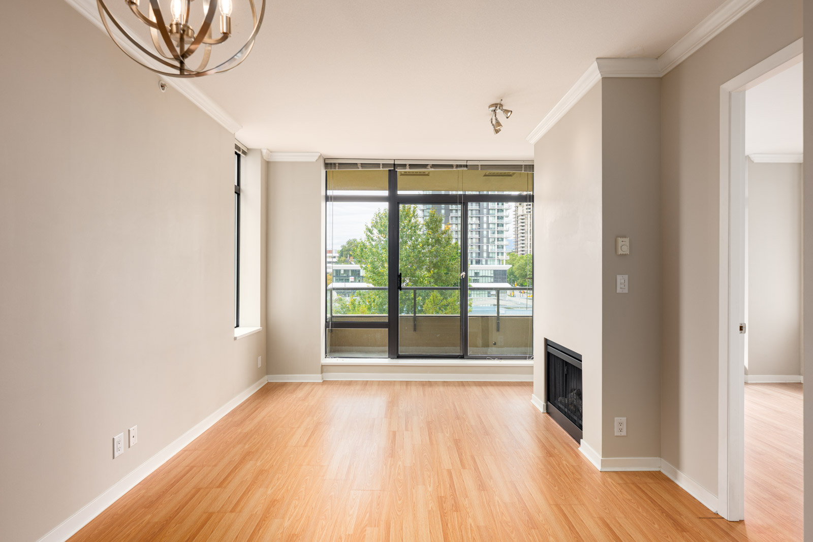 Unfurnished living room with light wood flooring, fireplace, and large windows facing a balcony with a city view. Adjacent room visible through open doorway.