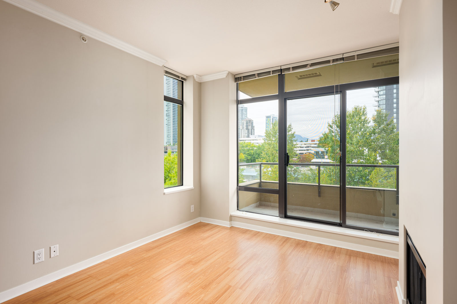 Unfurnished room with light wood flooring, beige walls, large window, and glass door leading to a balcony with a city and tree view.