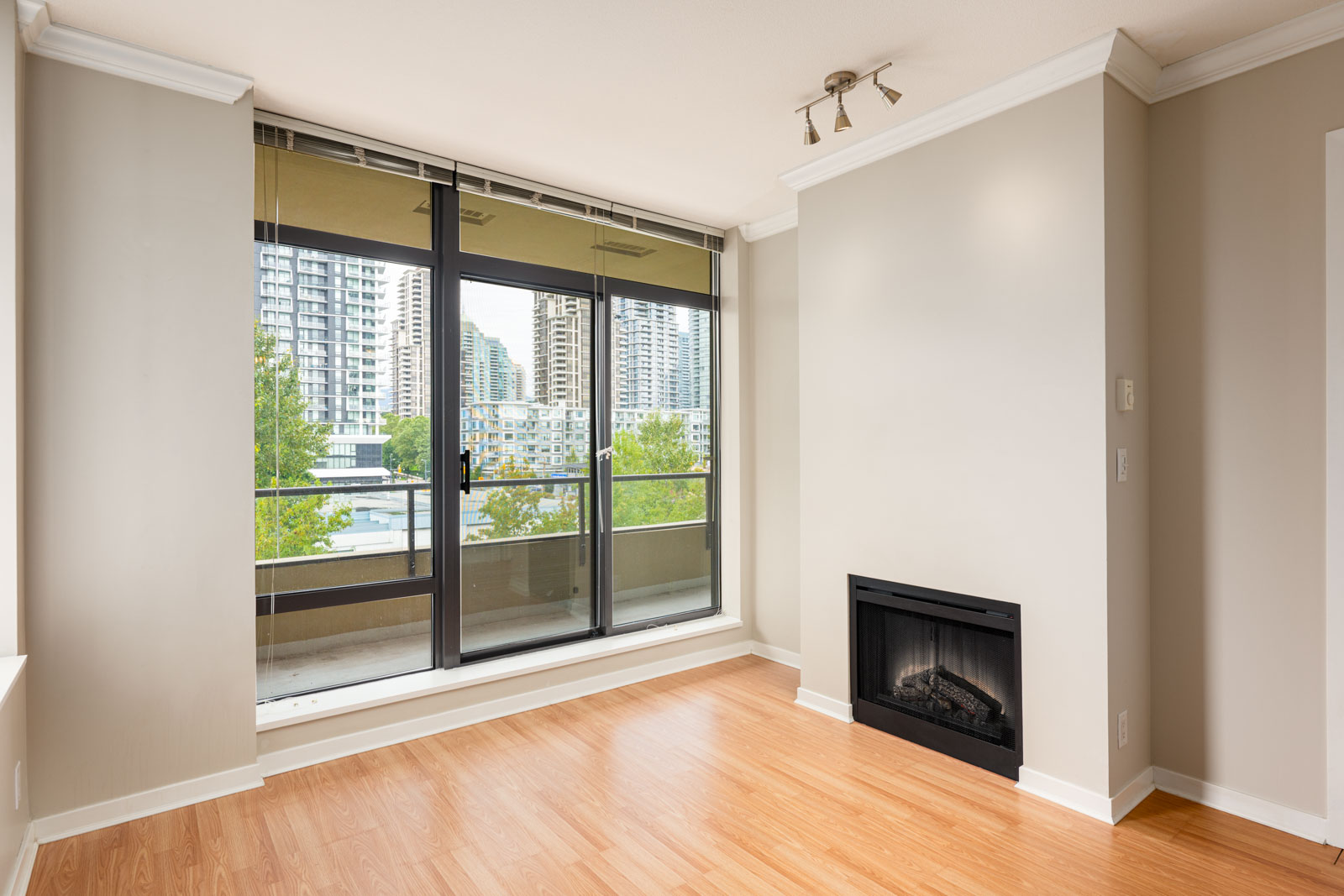 A modern empty room with light wood flooring, a wall-mounted fireplace, and large windows overlooking city buildings and greenery.