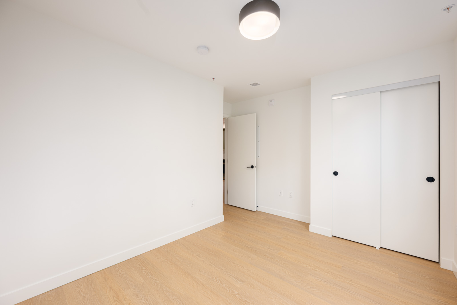 Empty room with light wood flooring, white walls, a closed closet with sliding doors, an open door, and a ceiling light fixture.