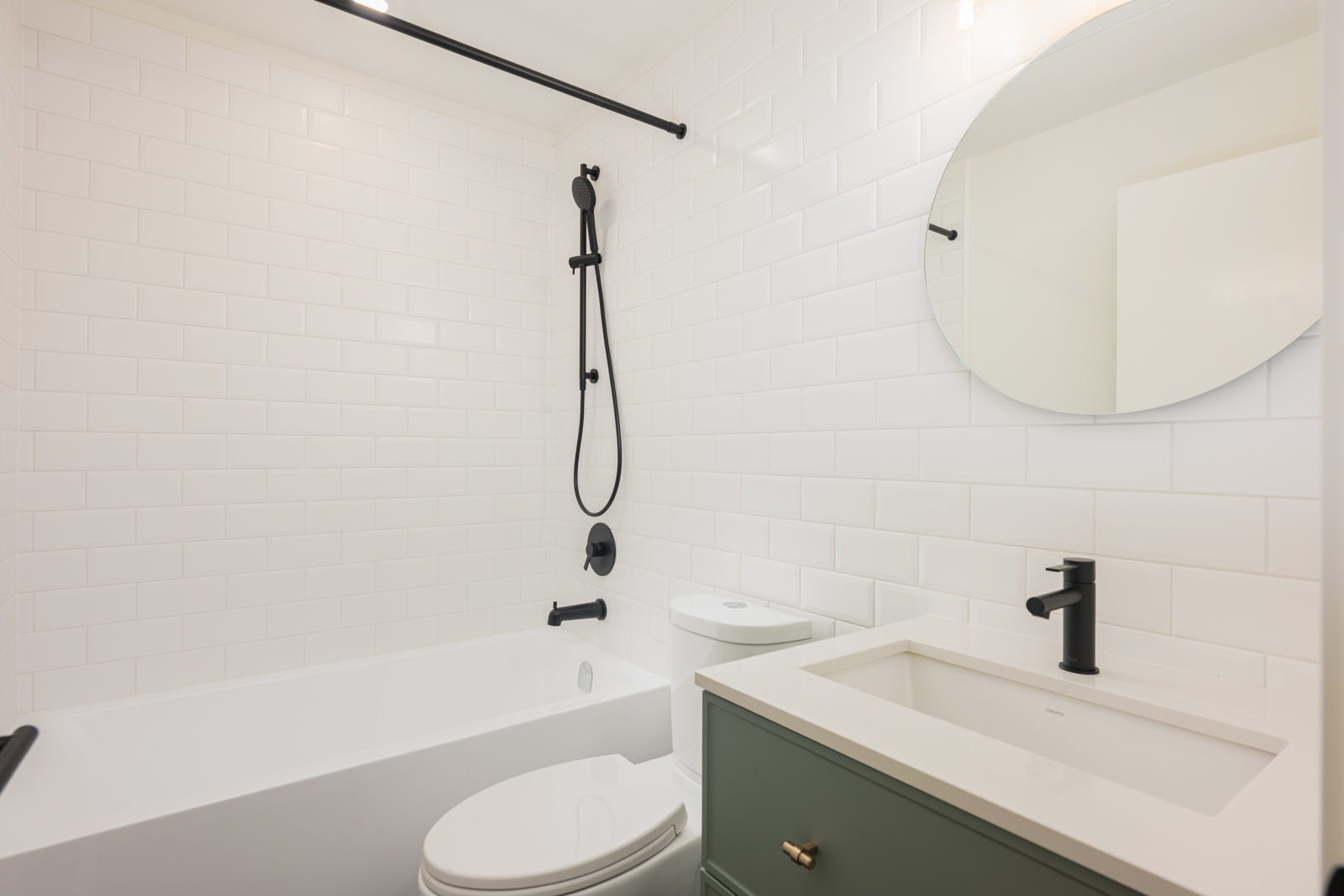 Modern bathroom with white subway tile, a bathtub with black fixtures, a round mirror, and a green vanity with a sink and black faucet.